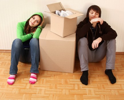 Two professional movers from Watson Removals London are inside a residential space, each wearing red shirts, blue overalls, and matching blue caps. The mover in the foreground is standing with arms crossed, smiling at the camera, next to a large cardboard box that is wrapped in clear plastic protective film. The second mover, positioned slightly behind and to the right, is also smiling and standing with one hand on his hip, near a cardboard moving box. The background features a white wall with a subtle, pale yellow abstract artwork, indicating a clean and modern interior setting. The scene suggests careful handling of household items, with the movers prepared to assist with a house removal in London or nearby areas, evident from the professional attire and organized environment. The lighting is bright, emphasizing clarity and professionalism, consistent with a typical house removal service environment used by Watson Removals London.