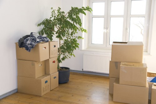 In a bright, spacious room with large windows letting in natural light, a professional removals specialist from Watson Removals London, dressed in red work overalls and a red cap, is seen handling packing materials, possibly preparing for a move within the London area. The specialist is pushing a small trolley loaded with wrapped household items, including a cushion and a bag, set against a backdrop of stacked cardboard moving boxes of various sizes. A young couple, standing nearby, appears engaged in the process; the woman, with long dark hair and casual clothing, is holding a small tablet and smiling, while the man, wearing a grey t-shirt and jeans, is holding a clipboard or document, possibly discussing the move. The environment suggests an interior residential space, likely an unoccupied living room or hallway, with minimal furnishings visible, focusing on the packing and moving process. Overall, the scene reflects professional house removal services in London, emphasizing careful handling of household goods, with a focus on organization and efficient relocation planning.