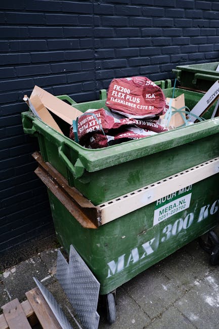 Two wooden boat-shaped recycling bins with open tops are positioned upright outdoors, each with a small wooden shelf inside, and are placed on a gravel surface. In front of each boat-shaped bin, there