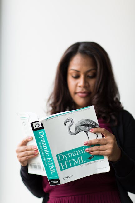A woman with dark hair is holding open a book titled 'Dynamic HTML' published by O'Reilly, displaying an illustration of a flamingo on a white page. She is indoors against a plain white background, we