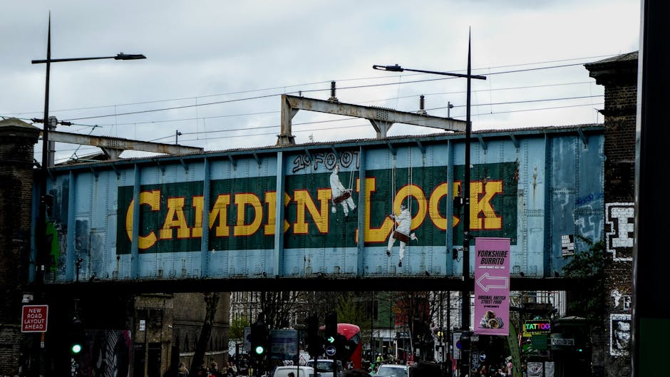 A large, weathered railway bridge painted with the words 'Camden Lock' in bold yellow and red lettering across a blue background, situated over a busy street in Camden, London. The bridge has visible