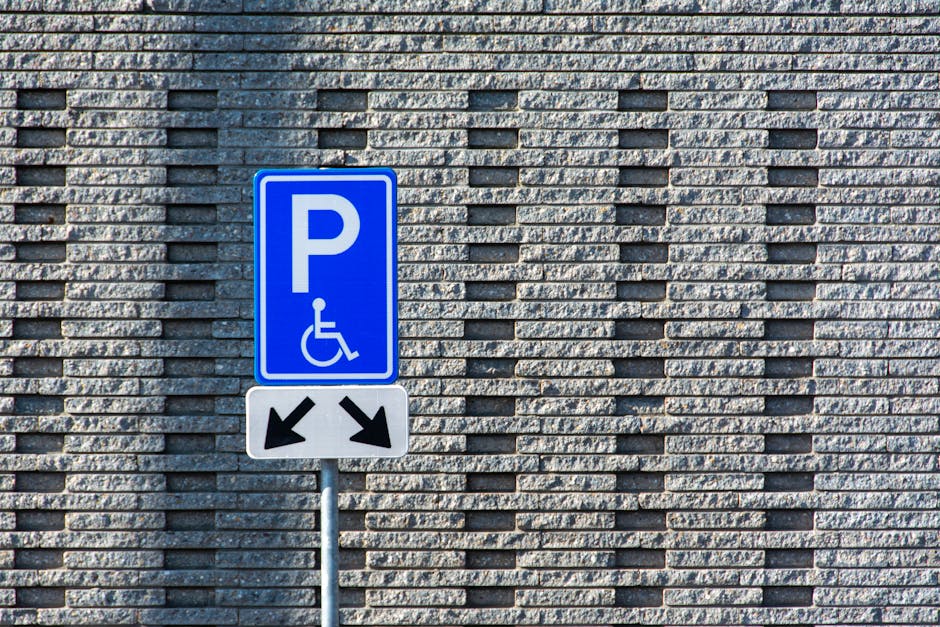 A blue parking sign with a white 'P' and a wheelchair symbol is mounted on a metal pole attached to a dark grey wall. The sign indicates designated accessible parking in Stratford. The shadow of the s
